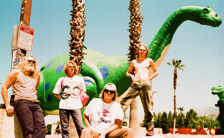 Australian psych-pop band Babe Rainbow posing outdoors near palm trees and a giant green dinosaur statue for a promotional band photo shoot.